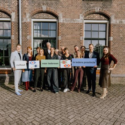 Een groepsfoto van de directeur-bestuurders van de provinciale ondersteuningsorganisaties. Ze houden bordjes vast met daarop het logo van de Raad van Twaalf en de zin ‘Cultuur voor iedereen in iedere provincie’.