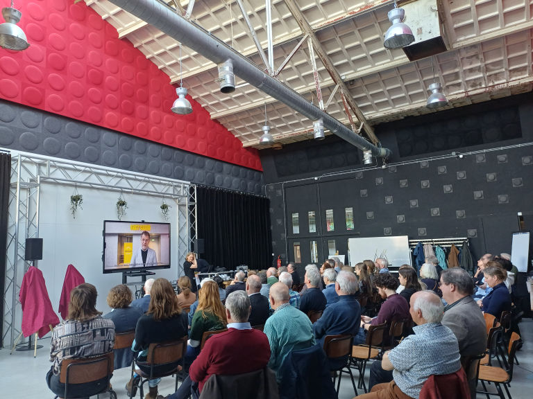 Mensen in een zaal in theateropstelling zitten en naar een presentatie kijken. De ruimte is licht, industrieel met een hoog plafond en rode en zwarte kleuren.