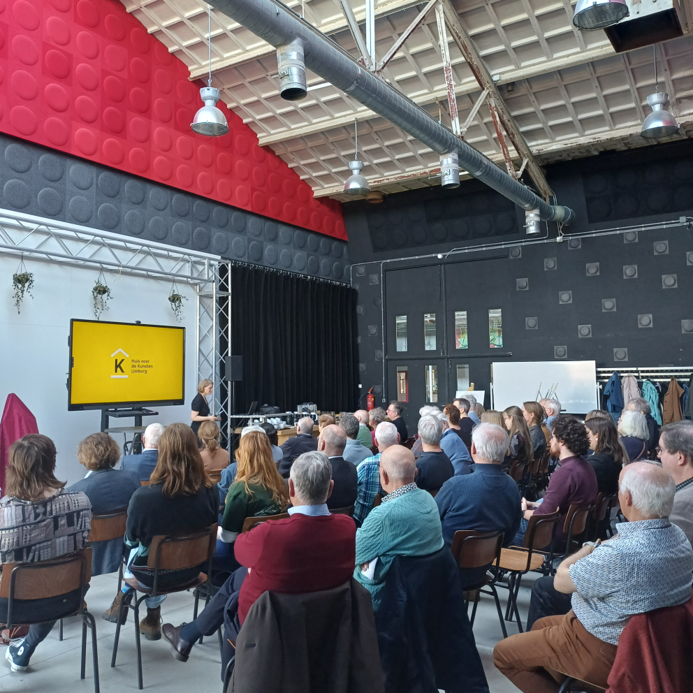 Mensen in een zaal in theateropstelling zitten en naar een presentatie kijken. De ruimte is licht, industrieel met een hoog plafond en rode en zwarte kleuren.