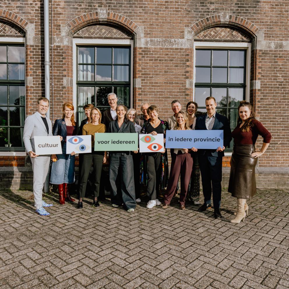 Een groepsfoto van de directeur-bestuurders van de provinciale ondersteuningsorganisaties. Ze houden bordjes vast met daarop het logo van de Raad van Twaalf en de zin ‘Cultuur voor iedereen in iedere provincie’.